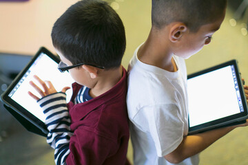 Two cute little boys stand back-to-back with their tablets during the break of their digital interactive class.