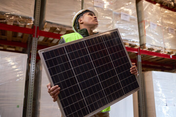 Low angle portrait of female worker carrying solar panel at indoor warehouse, copy space