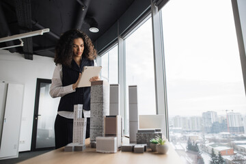 People, global business, company worker concept. Portrait of smiling lady with curly hair in formal clothes using tablet, posing in front of building complex prototype project of 3d city model.