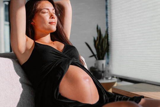 Pregnant woman relaxing sunbathing belly baby bump at home in sofa. Expectant mom tanning being happy in the sun indoors. Skincare, Vitamin D and healthy pregnancy concept image