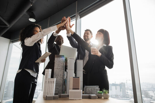 Portrait of team of positive successful multiethnic corporate coworkers at the office meeting, standing near table with 3d city model and giving high five as a sign fruitful collaboration.