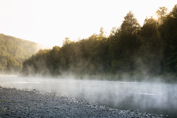 Idyllic gentle morning summer landscape with fog on river with white  haze flow above water, pebble shore, silhouette green forest on slopes in bright golden sunlight. Amazing misty wild nature.