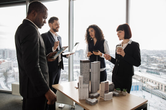 Multiethnic real estate agents, standing near table with 3d city model in office with panoramic city view, while handsome male project manager presenting them report about company's activity on laptop