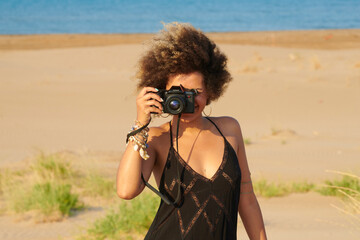 Portrait of smiling woman with dyed dark frizzy hair and black summer dress taking a photo of the photographer with professional camera. She is on the beach against the sea.