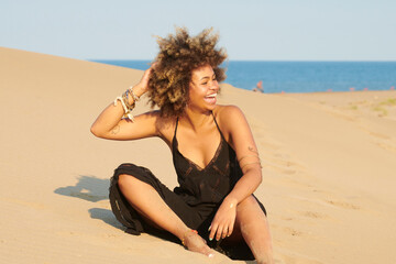 Portrait of delighted young woman with frizzy hair wearing black summer dress laughing on sandy dune against the sea holding arm behind her head.