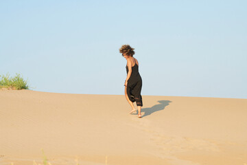 Content woman walking on sand. Ethnic woman in black dress strolling alone on sandy shore in bright sunshine