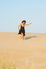 Full length portrait of cheerful woman with dark brown hair and black long summer dress running down the sandy hill happily with arm up. She is holding the skirt of dress.