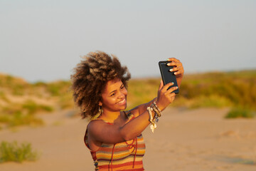 Lovely woman taking a selfie at sunset. Portrait of beautiful jovial woman with windy frizzy hair in printed bright top making selfie with her cell phone standing in sunlight at sundown