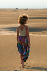 Stylish ethnic woman walking on sand. Back view of stylish modern black woman in colorful outfit walking barefoot on desert sand in sunlight 