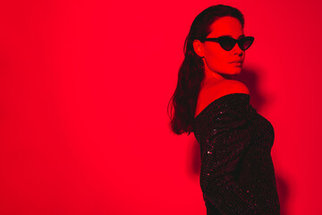Young beautiful confident brunette female in trendy evening shiny green reflecting dress. Sexy carefree woman isolated on red neon light in studio. Fashionable model in sunglasses. high fashion