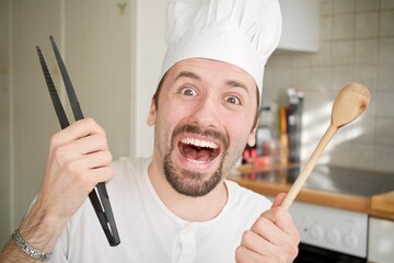 Happy chef in a professional kitchen, holding various cooking utensils. The well-organized...