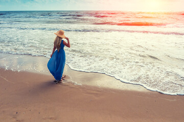 Seascape during sunrise. Woman on the beach. Young happy woman in a long fluttering blue dress and straw hat walks on the seashore