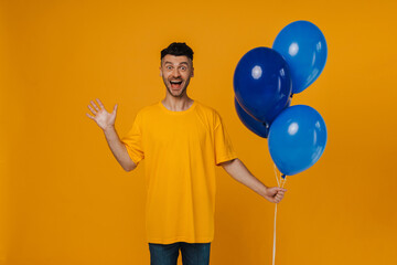 Cheerful man with blue air balloons waving hand isolated over yellow wall