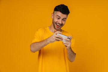 Joyful man playing online game on mobile phone isolated over yellow wall