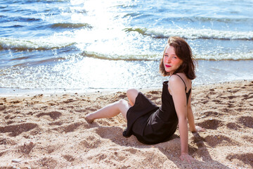 Winsome Happy Caucasian Brunette Girl in Casual Clothing Relaxing on Sandy Beach In Front of Sea Shore At Daytime.