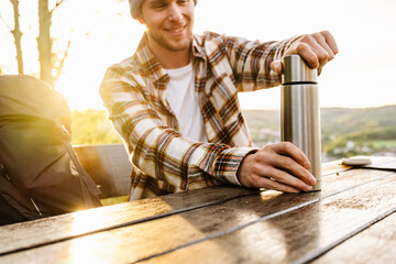 Smiling man tourist opening thermos while sitting on bench on hillside