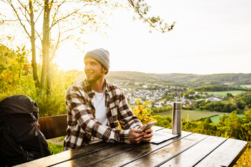 Fototapeta premium Young man tourist holding mobile phone and looking away while sitting on hillside