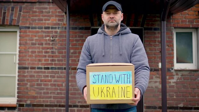 Responsible man volunteer holds cardboard box with donated food and humanitarian aids for ukrainian refugees leving from caring people in front of their house door. Help Ukraine in war with russian