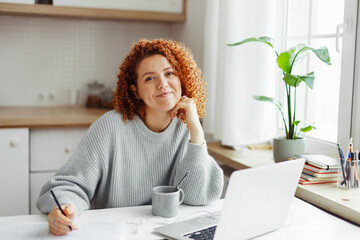 Cute curly redhead PR-manager of online store creating content plan working online at home, sitting at kitchen table with laptop and coffee cup, writing in copybook, making notes, looking at camera - 573477306