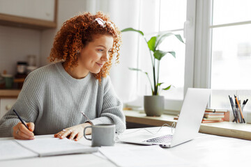 Cute university student with curly red hair doing homework sitting at kitchen table with coffee cup and wired earphones next to big window, watching online tutorial on laptop and writing in copybook - obrazy, fototapety, plakaty