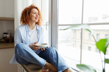 Young woman with red curly hair in jeans and shirt sitting on window sill looking through big...