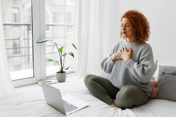 Pretty redhead plus size female sitting on bed in lotus posture with hands on chest doing pranayama techniques in front of laptop, breathing deeply, reaching inner balance after stressful day - obrazy, fototapety, plakaty