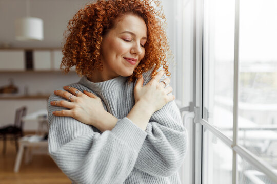 Horizontal image of pretty redhead female with closed eyes wearing rings on fingers, hugging herself, touching new soft sweater, enjoying comfort of fabric, standing next to panoramic window at home