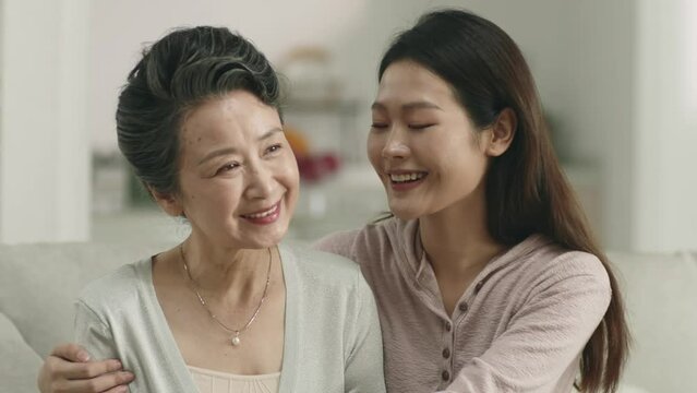 young asian adult daughter and senior mother sitting on couch relaxing in living room at home