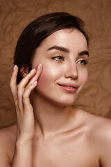 Well-kept skin. Beautiful young brunette girl taking care after skin, applying face moisturizer over brown studio background. Concept of natural beauty, skincare, cosmetology, plastic surgery, health