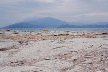 Panoramic Lake Garda view  from natural beach with white rocky shores at the end of city, overlooking crystal-clear waters and Mountains. Peninsula of Sirmione, Lombardy, Italy. Beautiful morning sky
