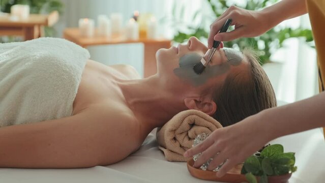 Cropped shot of unrecognizable beautician applying clay mask on beautiful face of young Caucasian woman lying on her back on lounger at luxury spa salon