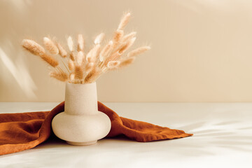 Ceramic vase with lagurus grass and brown linen towel at the background with warm shadows, bohemian...