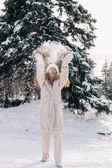 Young woman dressed in a light fur coat plays with snow in winter