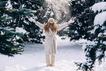 Young woman dressed in a light fur coat plays with snow in winter