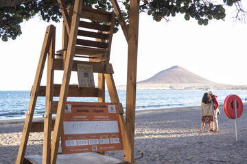 Lifeguard stand chair, under a leafy tree, on a sandy beach with ocean and mountain in the background. Calm ocean. Red Mountain. El Medano, Tenerife, Canary Islands, Spain. 