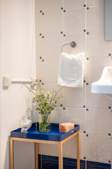 Corner of a bathroom with a decorative low decorative table with a blue base. On the table is a vase with wild flowers. On a tiled wall is a holder with a white terry towel.