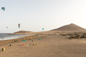 Obraz premium seascape, views of the yellow sand beach, bushes, people practicing kite surfing and Montaña Roja in the background. Paya Grande, El Medano, Tenerife, Canary Islands. Spain