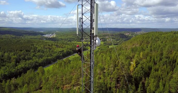 A high-angle view of a telecommunications technician working at height on a telecom tower, performing maintenance on 5G antenna.