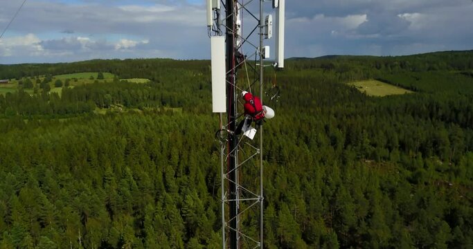 A high-angle view of a telecommunications technician working at height on a telecom tower, performing maintenance on 5G antenna.