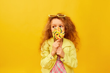 Hiding behind lollipop. Little cute girl, child with curly hair posing in bright clothes over yellow studio background. Concept of childhood, emotions, fun, fashion, lifestyle, facial expression