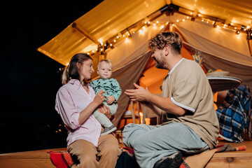 Fototapeta na wymiar Happy family with lovely baby relaxing and spend time together in glamping on summer evening near cozy bonfire. Luxury camping tent for outdoor recreation and recreation. Lifestyle concept