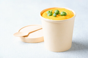 Creamy pumpkin fall soup in craft paper food container against light gray background. Butternut squash cream soup garnished with seasonings. Food delivery and soup to go. Mockup