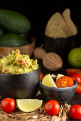 View of jar with guacamole on table with tomatoes, limes, avocados and nachos, black background, vertical