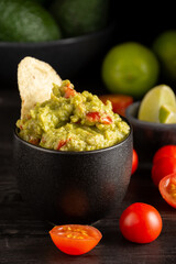 Close-up of black jar with guacamole on dark table with tomatoes, limes and avocados, black background, vertical