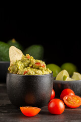 Close up of guacamole on dark table with tomatoes, limes and avocados, black background, vertical, with copy space