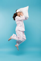 full length of barefoot young woman in night mask and sleepwear holding pillow and levitating on...