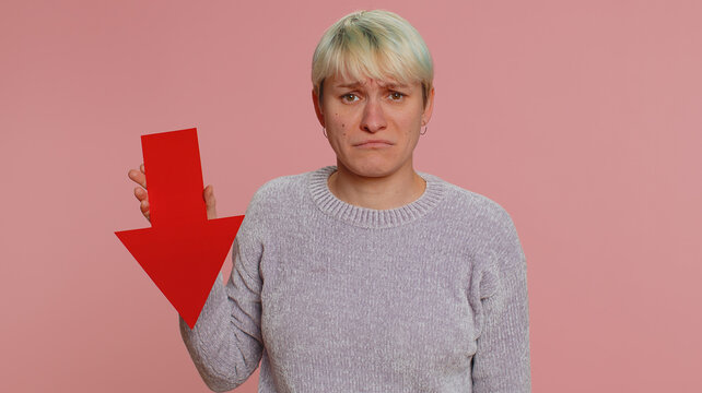 Upset young woman showing red arrow pointing down, concept of downgrade, unsuccessful business, fall of stock market money exchange rate, bankruptcy fail. Sad girl isolated on pink studio background