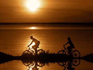 Fototapeta na wymiar sporty company friends on bicycles outdoors against sunset mountain. Silhouette of motion go of two 2 cyclist along shoreline coast Reflection sun on water. empty Copy Space for inscription.