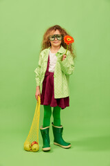 Autumn vibe. Little cute girl, child with curly hair posing with flower and fruits over green studio background. Concept of childhood, emotions, fun, fashion, lifestyle, facial expression