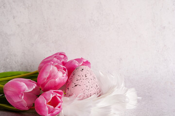 Easter egg in white nest of feathers with pink tulips on white concrete background